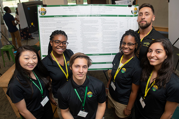 A group of six students in black collared shirts stand together in front of a poster board.