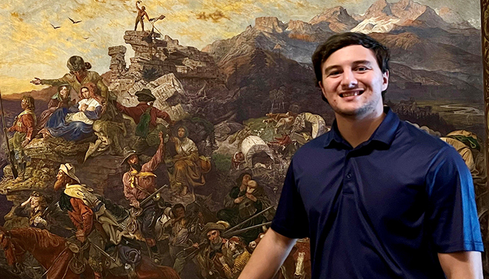 A young man in a blue shirt smiles and poses in front of a large painting of American pioneers.