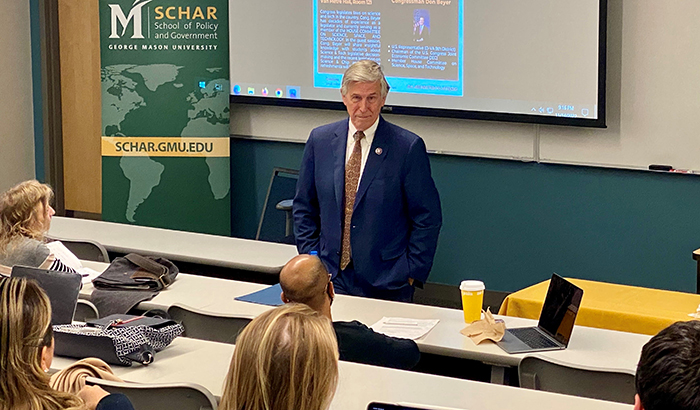 A man in blue suit stands in front of a Schar School banner while talking with students.