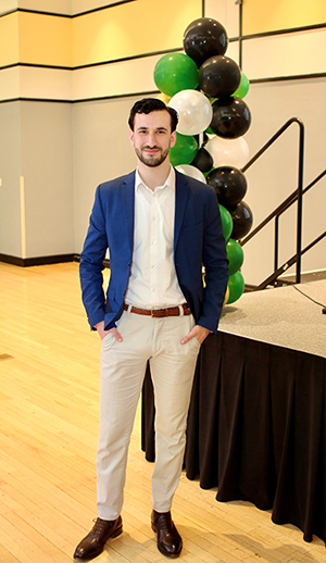 A man with a dark beard and jacket stands in front of balloons.