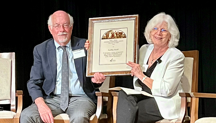 A man in a suit and a woman in white sit on a stage sharing a certificate.