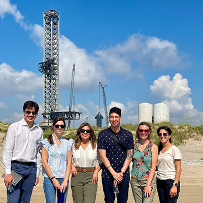 Six people stand in bright sunshine with a rocket launch pad behind them.