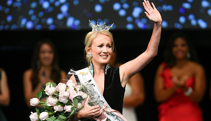 A woman in a black gown, holding roses, and wearing a silver crown cries as she waves to the audience.