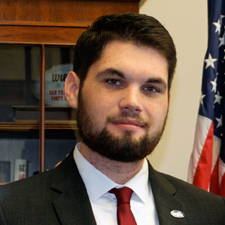 Greg Mercer, BA Government and International Politics major, stands with a black suit, white collared shirt, and red tie with an American flag in the background on the right.