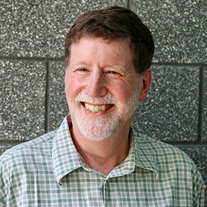 Mark N. Katz wearing a collared shirt stands smiling in front of a wall.