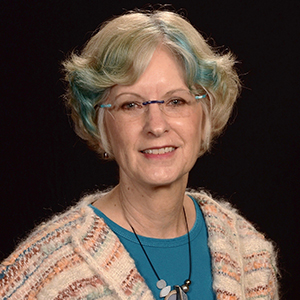 A woman with eyeglasses and a blue streak in her silver hair gazes at the camera.
