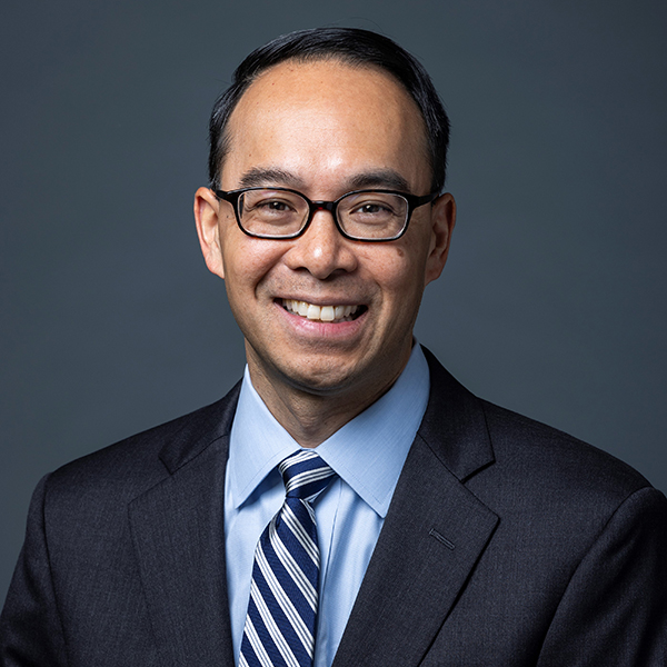 A man in a black suit, blue collared shirt, and a blue and silver striped tie stands and smiles