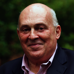 A man in a dark jacket and pink shirt smiles at the camera.