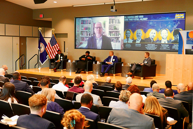 A wide-angle view of an auditorium shows four people seated on a stage in front of a seated audience.