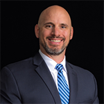 A man in a black suit, white collared shirt, and blue and white striped tie stands and smiles