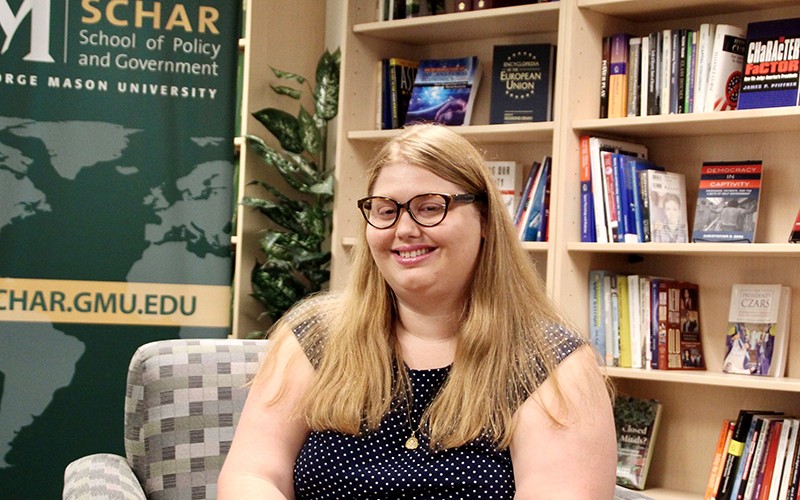 A woman in eyeglasses and a polka-dot top sits in front of book shelves.