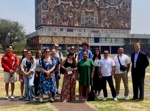 A group of people stand in front of a large and colorful mural in Mexico.