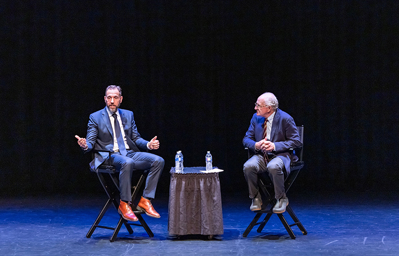 Two men sit in chairs on a stage