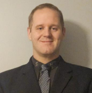 A man in a black blazer, black collared shirt, and gray tie stands and smiles.