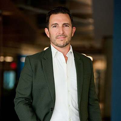 A man in a black blazer and white collared shirt stands and smiles