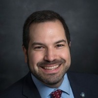 A man in a dark jacket and blue shirt smiles at the camera.