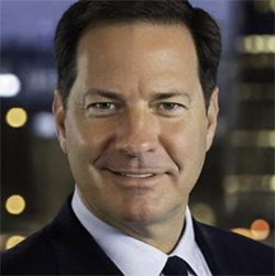 A man in a black suit, white collared shirt, and black tie, stands and smiles.