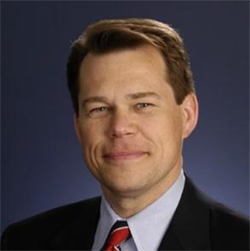A man in a black suite, white collared shirt, and red tie stands and smiles.