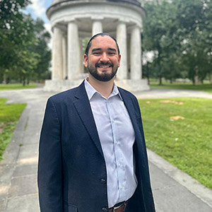 A man with a beard, blue blazer, and light blue collared shirt stands outside and smiles.