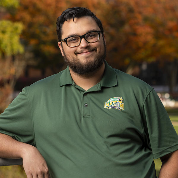 Zayd Hamid stands outside while smiling and wearing a green George Mason University collared shirt