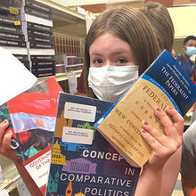A young woman in a mask holds up several books.