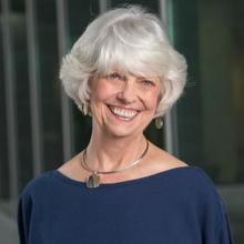 A woman with white hair and a blue shirt stands outside and smiles.
