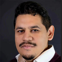 A man with a white collared shirt stands and smiles.