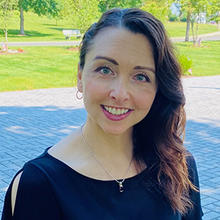 A woman with long brown hair and a black shirt stands outside and smiles.