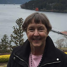 A woman in a black jacket and pink shirt stands outside and smiles.