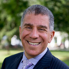 A man in a navy blue suit, blue collared shirt and a tie stands outside and smiles.