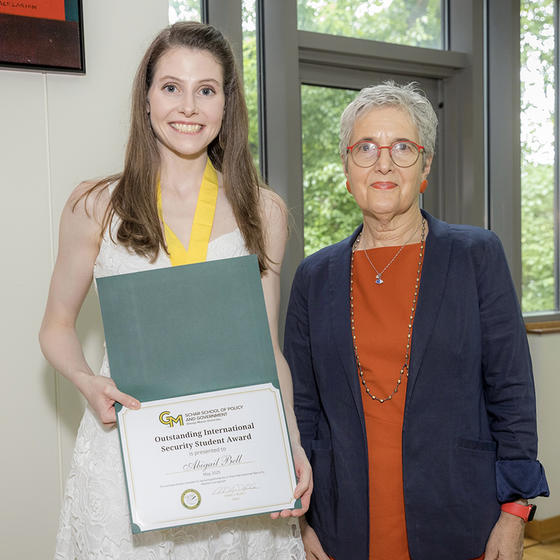 A woman in a white dress holding a certificate stands next to a woman in a blue jacket and red dress.