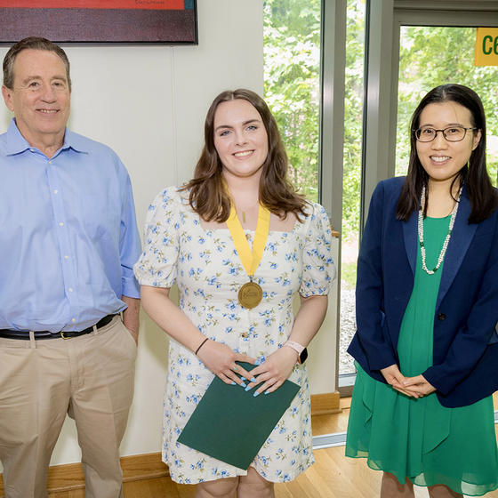 A man in a blue shirt stands next to a woman wearing a medal and another woman in a green dress and blue jacket.