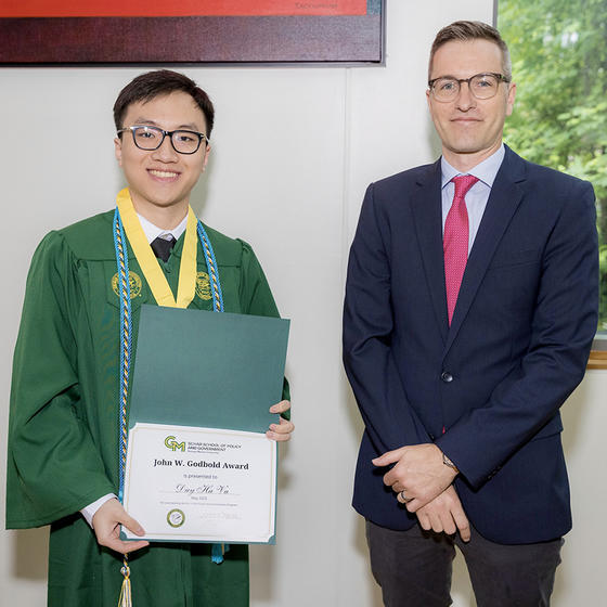 A man in a green gown holding a certificate stands next to a man in a blue suit.