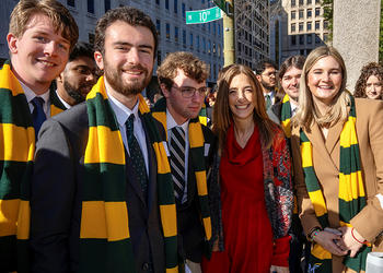 A woman in red stands amid several young people in green and gold winter scarfs on a street.