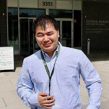 A young man in a blue striped shirt wearing a green lanyard stands in front of a building.