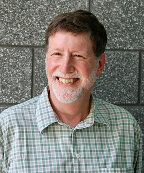 A man in a green checked shirt and a short white beard smiles off camera.
