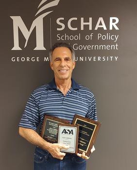 A man in a striped shirt poses under a Schar School sign and holds three awards.