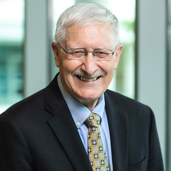 A man with silver hair wearing a dark jacket, blue shirt, and gold necktie smiles at the camera.