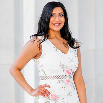 A young woman with long, dark hair stands in front of several marble pillars.