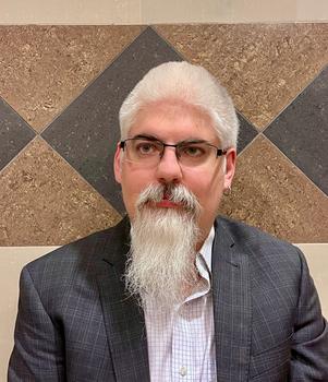 A man with white hair and a pointy white beard and eyeglasses sits in front of a geometric tile wall.