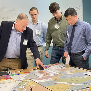 A man wearing a navy blazer points to a board featuring various countries while three  young men wearing name tags look on.