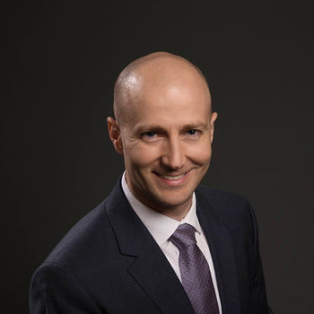 A bald man in a dark suit and tie leans toward the camera and smiles.