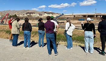 A group of people stand with their backs facing the camera in bright sunshine under a blue sky with white clouds.