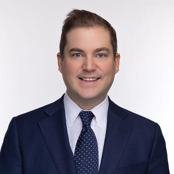 A man in a dark jacket and dark tie smiles at the camera.