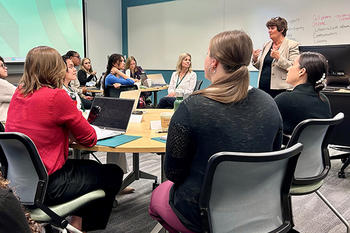 Women sit at tables in a classroom while a woman standing gestures with her hands.
