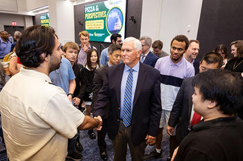 A man with white hair in a dark jacket shakes hands with a man while surrounded by others who are smiling.