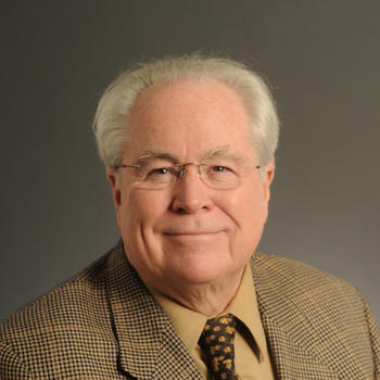 A man with white hair, eyeglasses, and wearing a tan plaid jacket gazes at the camera.