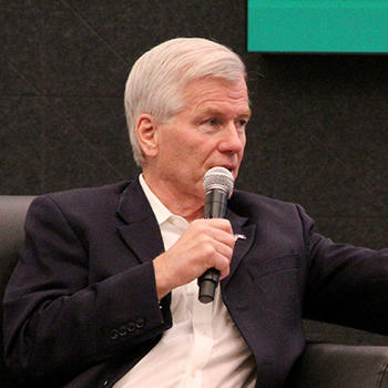 A man with white hair and holding a microphone gestures with his hand.