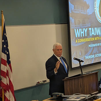 Former Vice President Mike Pence stands behind a podium beside the U.S. flag, addressing the audience during the “Why Taiwan Matters” event at George Mason University’s Schar School. A presentation slide with the event title is displayed behind him.