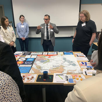 An instructor addresses a circle of students gathered around a tabletop simulation map of the South China Sea during a Schar School crisis simulation.